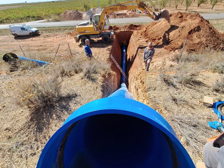 Tubería principal de riego de gran diámetro colocada en zanja junto a una parcela de viñedo y almendro, con retroexcavadora al fondo